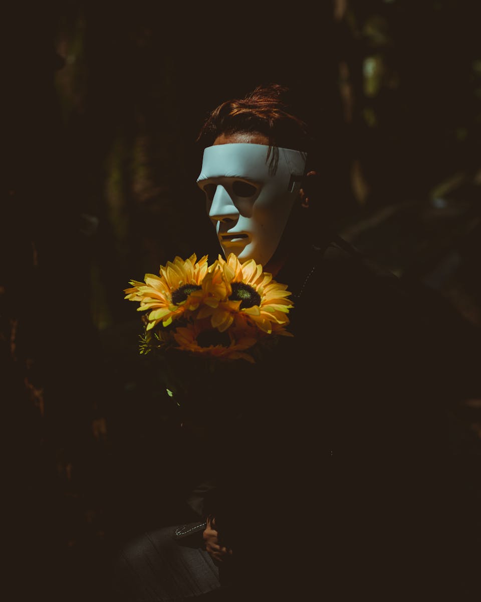 A person wearing a mask holds sunflowers, casting a mysterious and eerie vibe in dim lighting.