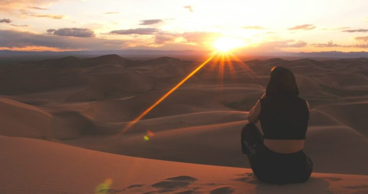 woman in black jacket sitting on sand during sunset